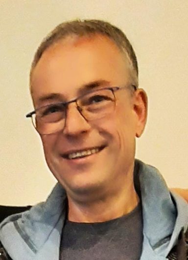 Ein lachender Mann mit Brille und kurzen grauen Haaren, trägt ein graues Oberteil.