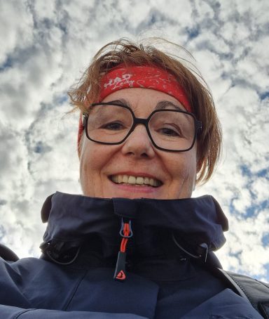 Lächelnde Person mit Brille und rotem Stirnband vor bewölktem Himmel.