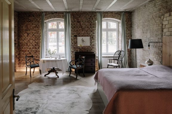Helles Schlafzimmer mit Ziegelwänden, Bett, Sesseln und Fenster mit Vorhängen.