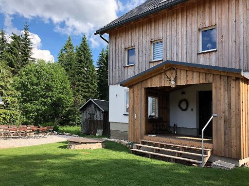 Holzhaus mit Terrasse, umgeben von Bäumen und einer kleinen Feuerschale im Garten.