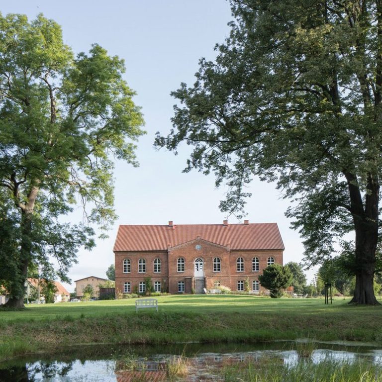 Zweistöckiges, rotes Backsteingebäude mit großen Fenstern und einem Garten davor.